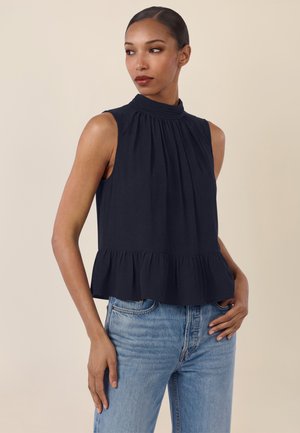 Woman with dark hair in a high-neck, sleeveless black top with ruffled hem and light blue jeans, standing against a beige background.