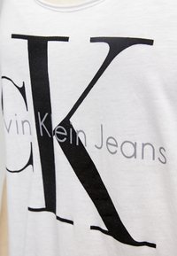Bílý bavlněný tílko se širokým černým nápisem "CK" a "Calvin Klein Jeans" v šedé, s kulatým výstřihem a bez rukávů.