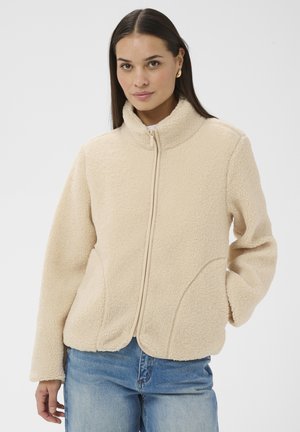 Frau trägt eine beige Fleecejacke mit Reißverschluss und Taschen sowie blaue Jeans, steht vor einem schlichten hellen Hintergrund.