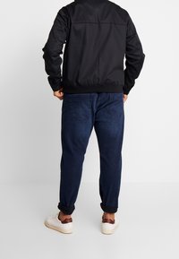 Man i svart jacka, mörkblå upprullade jeans och vita sneakers med vinröd häl detalj, står framför en helt vit bakgrund.