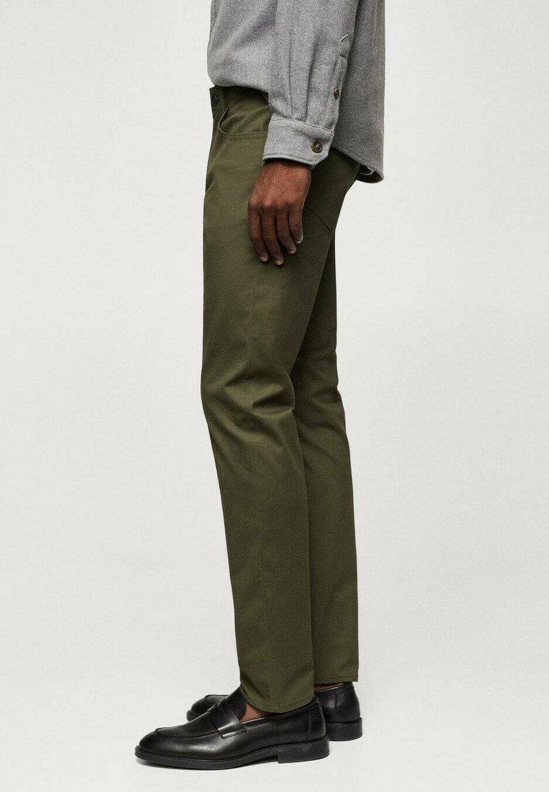 Khaki grüne maßgeschneiderte Hose mit geradem Schnitt, aus glattem Stoff. Kombiniert mit schwarzen Slippern und einem grauen Hemd mit Knopf.