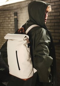 White roll-top backpack with brown leather accents, a front zip pocket, and adjustable straps, set against a textured brick background.