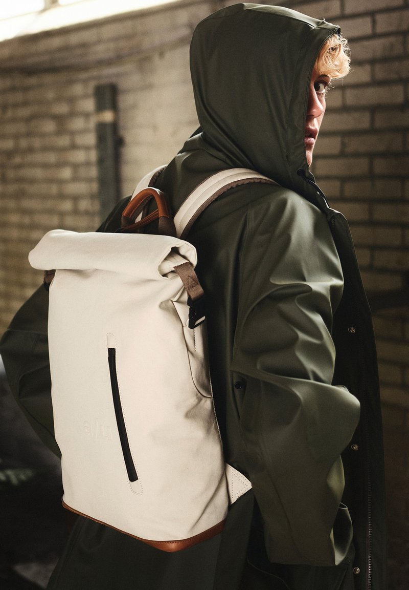 White roll-top backpack with brown leather accents, a front zip pocket, and adjustable straps, set against a textured brick background.