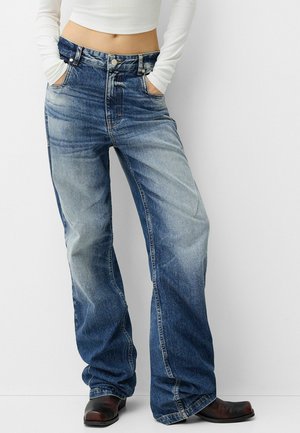 Osoba nosząca wyblakłe, luźne niebieskie dżinsy z rękami w kieszeniach, w połączeniu z białym, długim, krótkim topem i czarnymi butami.