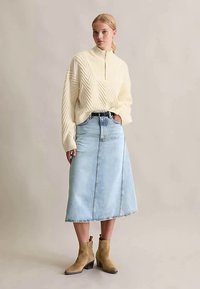 Kobieta w kremowym, fakturowanym swetrze z zamkiem błyskawicznym, jasnoniebieskiej spódnicy midi z denimu z czarnym paskiem i beżowych zamszowych botkach za kostkę, stojąca na tle jednolitym.