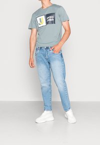 Jasnoniebieska graficzna koszulka z krótkimi rękawami, z czarno-żółtym logo "JACK & JONES", zestawiona z wyblakłymi niebieskimi dżinsami i białymi trampkami.