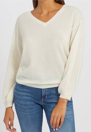 Pull à col V en crème, fait en tissu tricot doux. Il présente des manches longues, bouffantes et un ourlet côtelé. Associé à un jean en denim bleu.