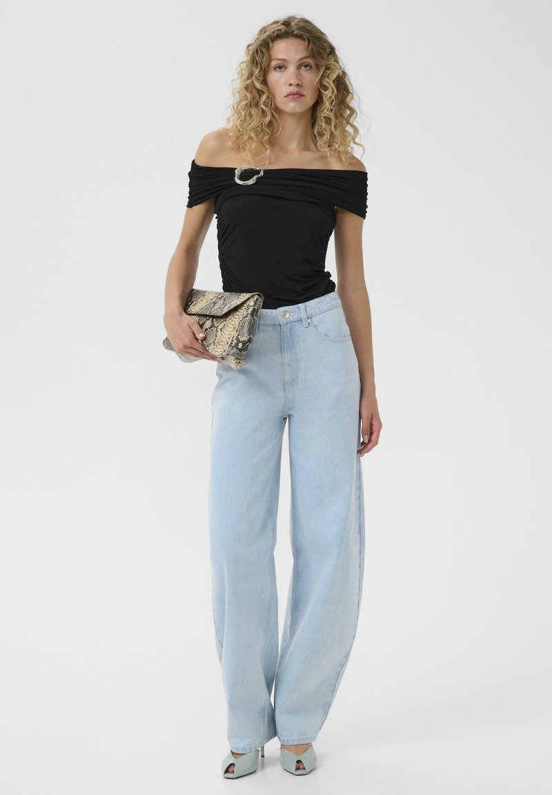 Vrouw met krullend haar die een zwart topje met blote schouders draagt, lichtblauwe wijde jeans, lichtblauwe hakken en een clutch van slangenleer vasthoudt.