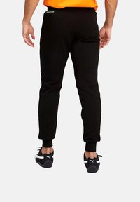 Pantalones jogger negros hechos de tela suave, con un diseño cónico, puños acanalados y un pequeño logo en la parte trasera. Combinados con zapatillas negras.