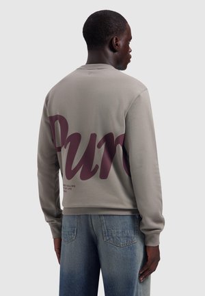 Man wearing light gray sweatshirt with large purple text and faded blue jeans, facing away against plain light background.