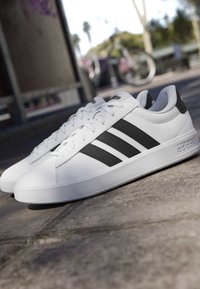 Baskets blanches Adidas avec des rayures noires posées sur une surface en béton, avec un vélo flou et un arrière-plan extérieur.