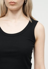 Νεαρή γυναίκα με ξανθά μαλλιά, φορώντας μαύρο ριμπ αμάνικο τοπ και ασημένια κρίκους σκουλαρίκια, μπροστά σε απλό φωτεινό φόντο.