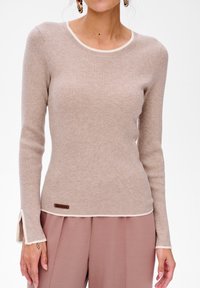Beige geribbelde langwerpige top met crèmekleurige afwerking bij de halslijn en manchet. Beschikt over een klein bruin label aan de zoom.
