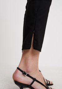 Černé přiléhavé kalhoty se stranním rozparkem na lemu, doplněné o černé sandály na podpatku se čtvercovou sponou. Hladká textura látky.