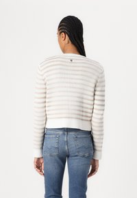 Gestreifter, gekürzter Pullover in Creme und hellem Beige, mit langen Ärmeln, geripptem Saum und kleinem Logo-Detail auf dem Rücken. Kombiniert mit blauen Jeans.