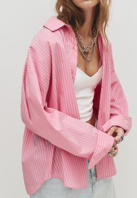 Camicia a maniche lunghe rosa chiaro con righe verticali bianche, caratterizzata da un fit ampio, colletto appuntito e chiusura a bottoni. Indossata sopra una canotta bianca.