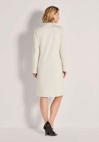 Frau, die mit dem Rücken zur Kamera steht, trägt ein knie-langes, langärmeliges cremefarbenes Kleid und schwarze High-Heels vor einem einheitlichen Hintergrund.