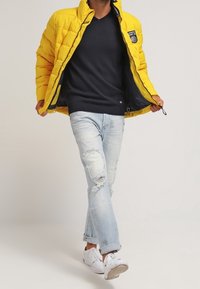 Doudoune jaune avec col à zip, pull à col en V bleu marine, jean bleu clair déchiré et baskets blanches, avec des patchs visibles aux genoux.