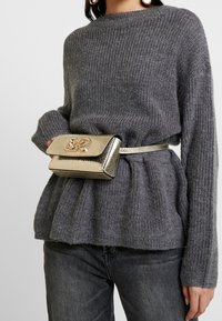 Harmaa ribattu neulepaita pitkilla hihoilla; kultainen metallinen vyölaukku, jossa on teksturoitu pinta ja koristeellinen soljettiedosto.