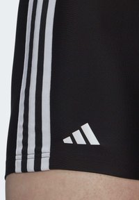 Pantalones cortos deportivos negros con tres franjas verticales blancas y un logo de Adidas blanco en el dobladillo. Textura de tela suave, diseño ajustado.