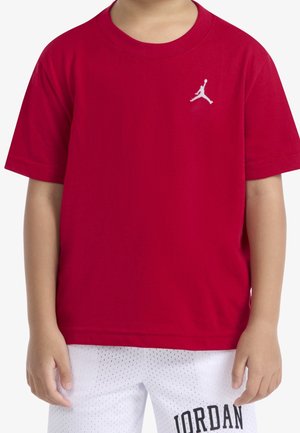 Enfant portant un t-shirt rouge avec un logo Jumpman blanc et un short blanc perforé avec le texte « JORDAN » sur la jambe gauche.