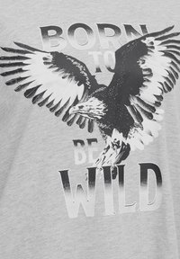 Camiseta gris con un gráfico en blanco y negro de un águila con las alas extendidas, superpuesto con el texto "NACIDO PARA SER SALVAJE."