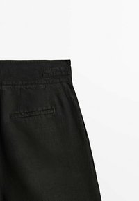 Gros plan d’un pantalon noir montrant la poche arrière et le détail de la ceinture sur fond blanc.