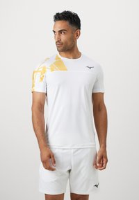 Witte sportshirt met korte mouwen en een geel geometrisch patroon op de schouder. Bevat een zwart logo op de borst.