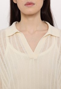 Strikket polo skjorte i lysekræm med lodrette striber. Har en V-halsudskæring, ribbet tekstur og afslappet krave for et åndbart design.