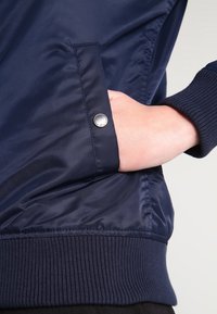 Námořnická modrá bomber bunda s hladkým textilním povrchem, žebrovanými manžetami a viditelnou kapsou se stříbrným patentním knoflíkem.