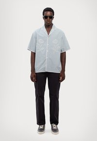 Šviesiai mėlyna trumpomis rankovėmis marškiniai su gėlių siuvinėjimu, balta mygtukų juosta, derinami su tamsiais kelnėmis ir pilkais sportbatiais, užsidėti saulės akiniai.