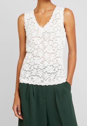 Haut en dentelle blanche sans manches à motif floral, col en V, porté rentré dans un pantalon plissé taille haute vert foncé.