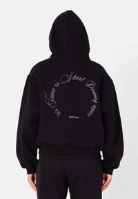 Zwarte hoodie met een cirkelvormige tekstgrafiek in het wit op de achterkant, met de frase "Het is tijd om te beginnen dromen" en logo-detail.