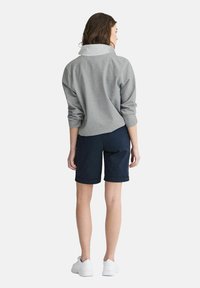 Grå fleece-pullover med vit krage, tillsammans med marinblå shorts. Modell som bär vita sneakers, sedd bakifrån.