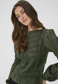 Junge Frau mit langen braunen Haaren, die eine strukturiert olivgrüne Bluse mit langen Ärmeln trägt, sanft lächelnd, vor einem einfarbigen hellen Hintergrund.