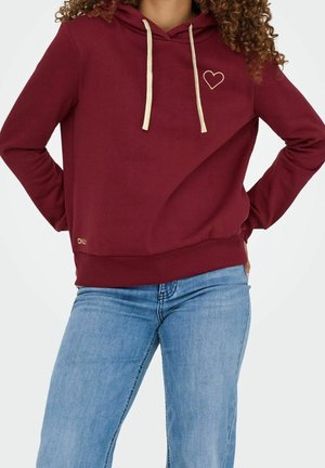 Woman wearing a burgundy hoodie with cream drawstrings and a small heart outline, paired with light blue jeans, hands on hips.