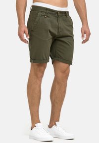 Shorts verts olive en coton, avec une coupe slim, des revers roulés, une fermeture à bouton, et des poches. Associés à des baskets blanches.