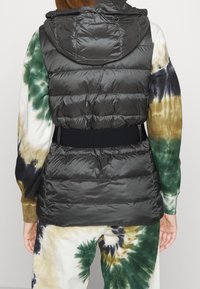 Vadderad svart väst med huva, med horisontell quiltad textur och ett svart elastiskt bälte i midjan, bärs över en tie-dye-outfit.