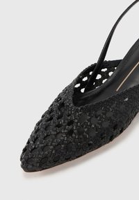Fekete szőtt bőr slingback cipő hegyes orral, bonyolult kivágott mintával és vékony pánttal, fehér háttér előtt.
