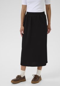 Jupe noire mi-longue avec une taille élastique, assortie à des chaussures marron et des chaussettes crème à la cheville. Tissu lisse et coupe décontractée.