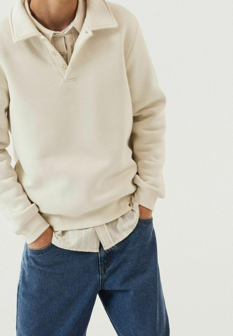Personne portant un pull crème par-dessus une chemise beige boutonnée, les mains dans les poches d'un jean bleu, debout devant un fond blanc uni.