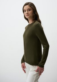 Maglione verde oliva realizzato in morbido tessuto a maglia, con maniche lunghe, scollo rotondo e polsini a coste, abbinato a pantaloni bianchi.