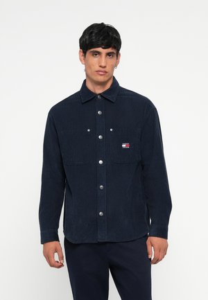 Navy corduroy shirt with button front, two chest pockets, and logo patch. Features a collared neckline and long sleeves. Textured surface.