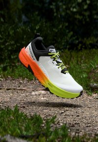 Παπούτσι για τρέξιμο σε μονοπάτι με λευκό επάνω μέρος, μαύρο γιακά και διαβαθμισμένο μεσαίο πέλμα από λαχανί πράσινο έως φωτεινό πορτοκαλί, αιωρούμενο πάνω από βραχώδη διαδρομή.
