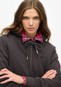 Sudadera gris oscuro con cordón y herrajes plateados, superpuesta sobre una camisa de cuadros roja y rosa, con logo de la marca bordado.