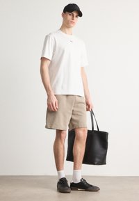 T-shirt bianco a maniche corte con logo, pantaloni corti beige, sneakers nere, calze bianche e una borsa tote nera. Design semplice e colori neutri.