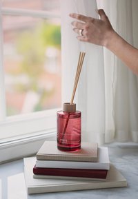 Diffuseur en verre rouge avec un bouchon en bois et des bâtonnets de rotin, posé sur des livres empilés de couleur neutre sur une surface en marbre près d'une fenêtre.