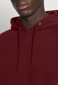 Felpa bordeaux con una texture morbida, dotata di cappuccio con coulisse, occhielli in metallo e polsini a costine. La vista ravvicinata evidenzia i dettagli delle cuciture.