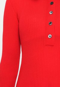 Haut en tricot rouge côtelé avec un plastron à boutons comportant quatre boutons noirs, des manches ajustées et un décolleté droit.