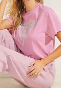 Frau trägt ein hellrosa Guess-T-Shirt und passende Hose, sitzt mit einem angezogenen Knie und der Hand auf dem Oberschenkel.